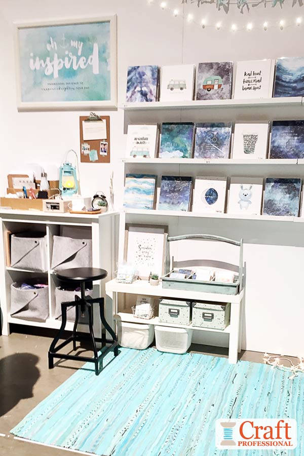 The booth sign painted in the same style as the art prints and cards for sale in this craft booth shows the artist's style from a distance. Click through for 6 more craft booth display ideas. Watercolor art prints and cards in shades of blue on display at a craft show. Large booth sign painted in the same style shows the artist's aesthetic.
