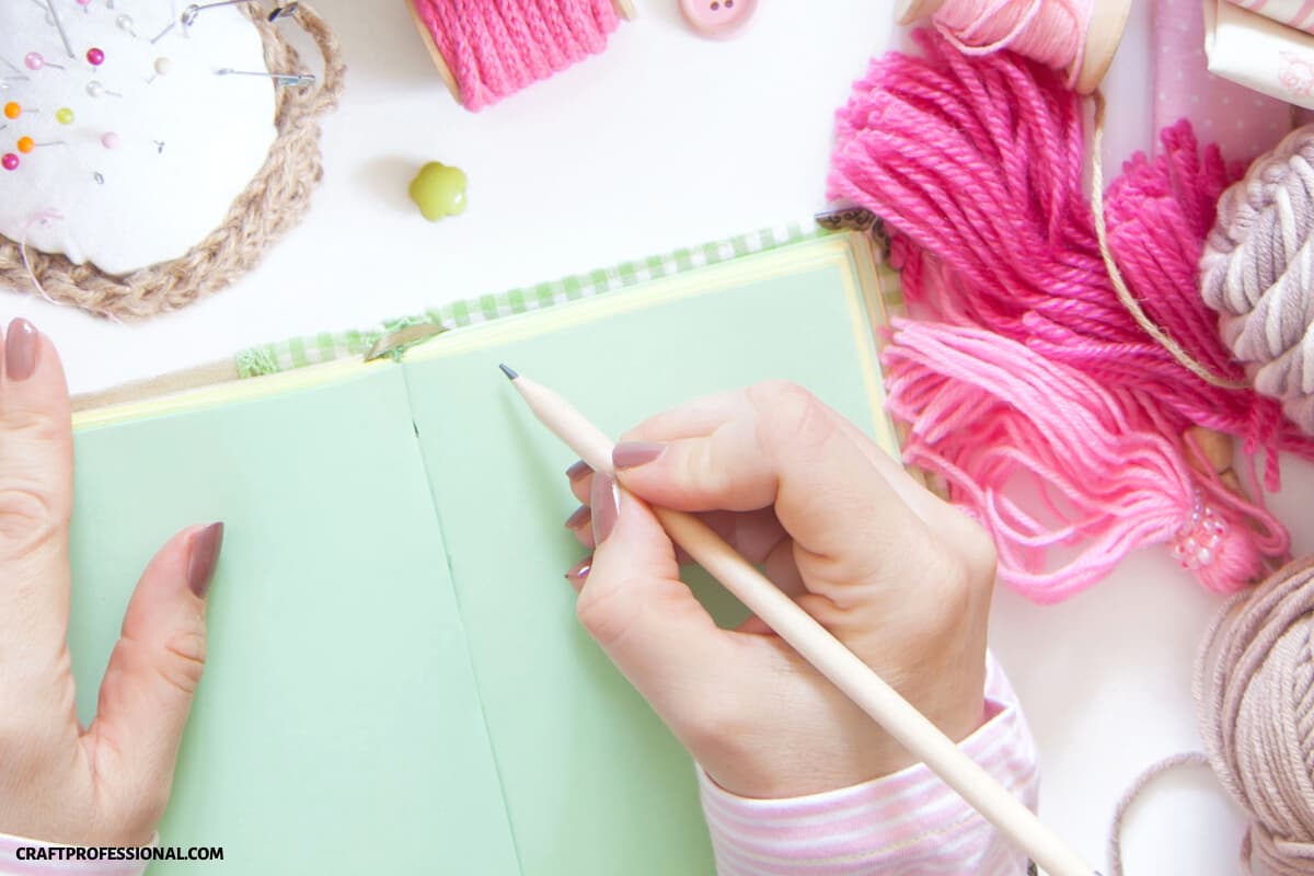 Woman writing in a notebook surrounded by yarn, thread, and a pincushion.