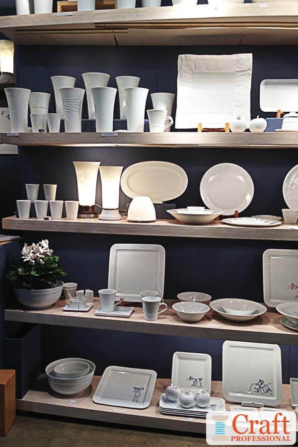White, handmade tableware displayed in a craft booth on rows of light wood shelves with a midnight blue backdrop, and plenty of lighting under each shelf. White, handmade tableware displayed in a craft booth on rows of light wood shelves with a midnight blue backdrop, and plenty of lighting under each shelf.