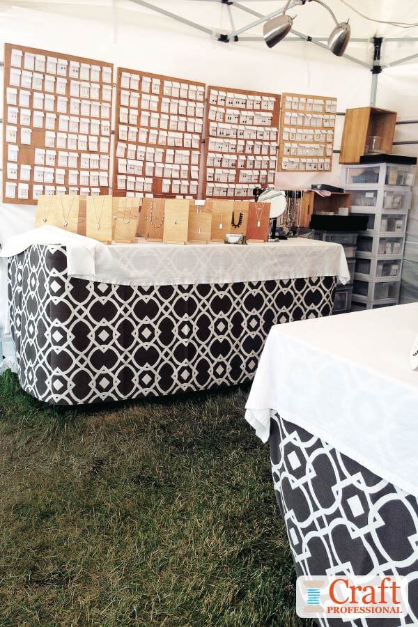 Well lit handmade jewelry displayed on shelves and tables at an outdoor craft show. Handmade jewelry displayed on tabletops and shelves at an outdoor craft show. Tables are covered in graphic print black and white tablecloths..