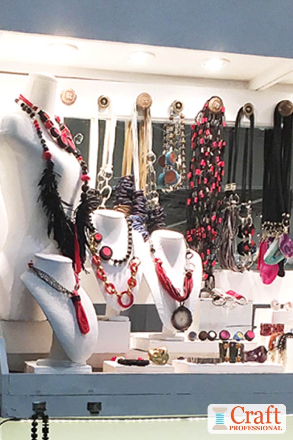 Handmade jewelry displayed on four white busts of varying sizes and arranged to form a pyramid shape. More jewelry on display in the background.