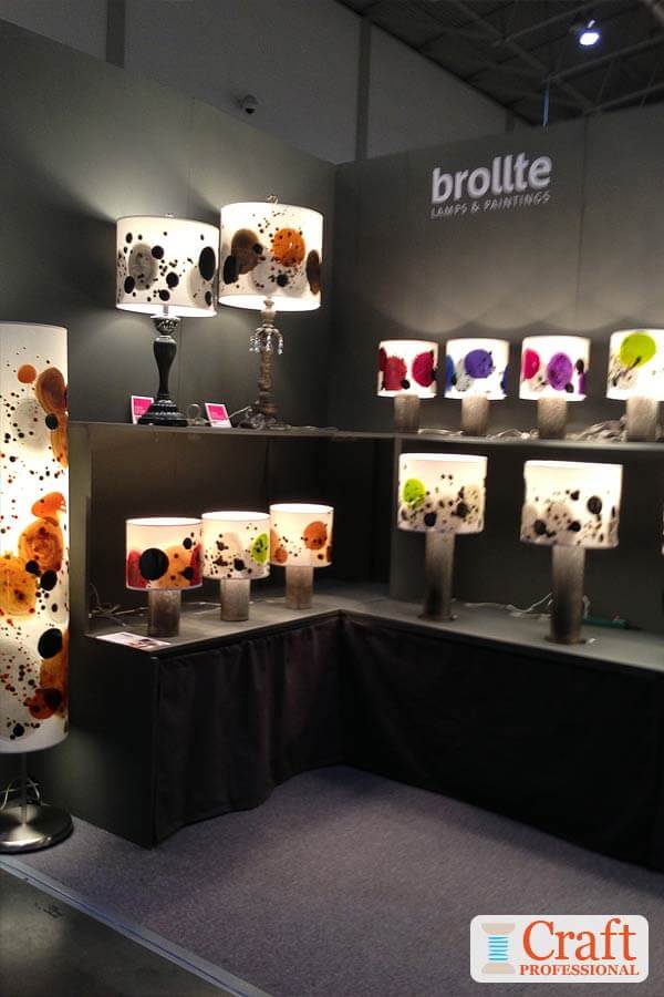 Handmade white lamps with colorful abstract designs are displayed against a black backdrop at a craft show. The lamps are turned on, providing their own illumination for the booth.