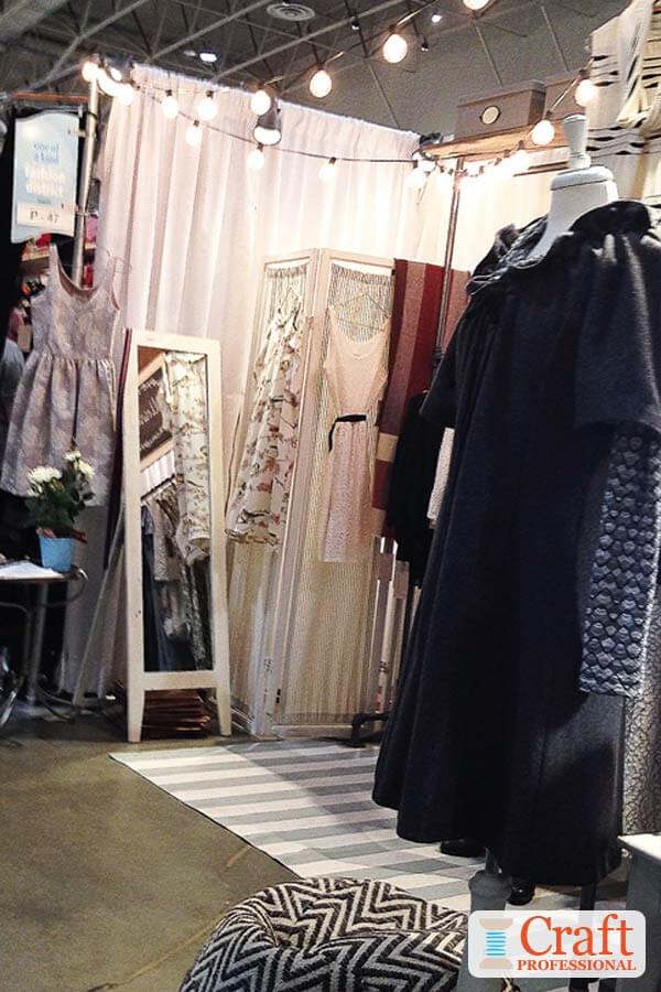 Handmade clothing displayed at a craft show. Strings of globe shaped lights are strung across the top, of the booth adding lighting and style to the display.