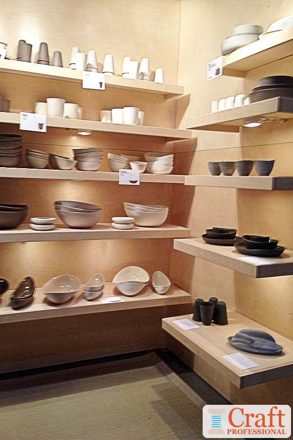 Neutral colored, handmade tableware displayed in a craft booth on rows of light wood shelves with lots of lighting under each shelf. Neutral colored, handmade tableware displayed in a craft booth on rows of light wood shelves with lots of lighting under each shelf.
