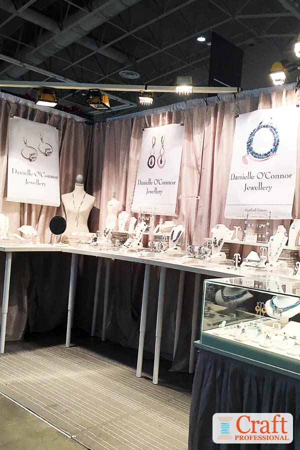 The three posters in this handmade jewelry booth with large-scale product photos help to draw shoppers' attention from a distance. Click through for 6 more merchandising ideas for craft show displays. Handmade jewelry on display on tables at a craft show. Three posters with large-scale photos of the jewelry help to draw shoppers' attention from a distance.